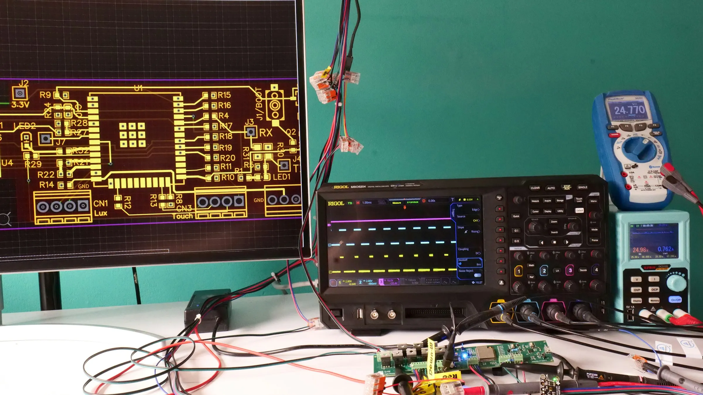 Elektronik-Arbeitsplatz mit Platinen-Layout am Monitor, Oszilloskop, Multimeter und Testplatinen